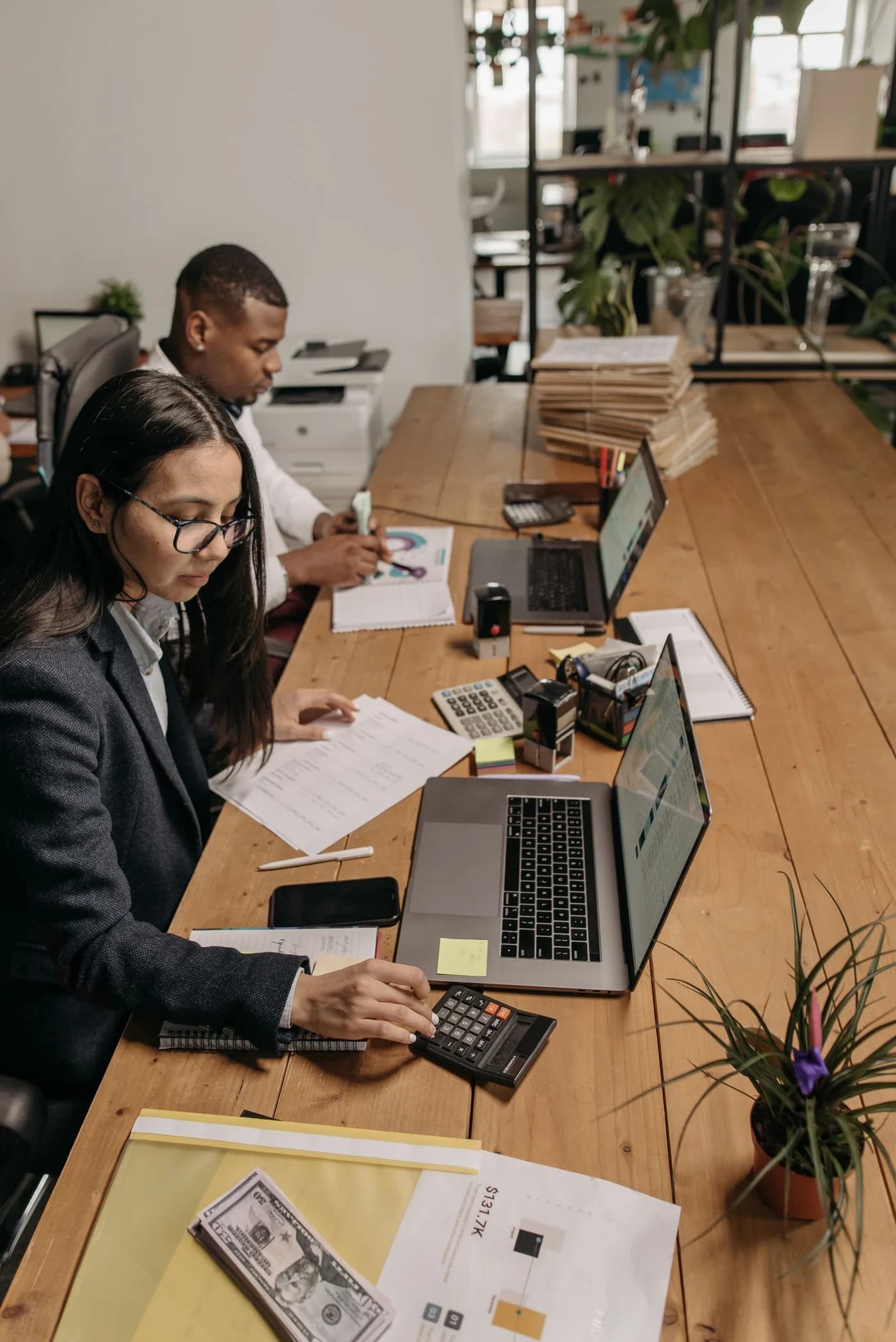 Two colleagues working with laptops and calculators at a shared desk.